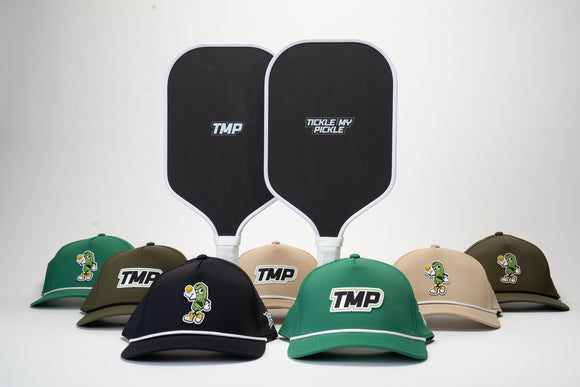 Two TMP Stealth Pickleball Paddles standing side by side, showing the TMP face and Tickle My Pickle face, with the TMP Performance Pickleball Hat lineup arranged at the base