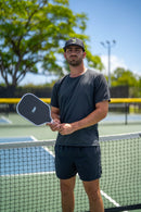 Player in a TMP cap holding the TMP Stealth Pickleball Paddle horizontally across his torso, with a pickleball court blurred in the background