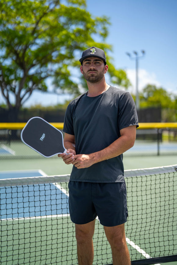 Player in a TMP cap holding the TMP Stealth Pickleball Paddle horizontally across his torso, with a pickleball court blurred in the background
