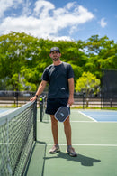 Player wearing a TMP cap and athletic gear standing next to a pickleball net, holding the TMP Stealth Pickleball Paddle by his side, trees and sky behind
