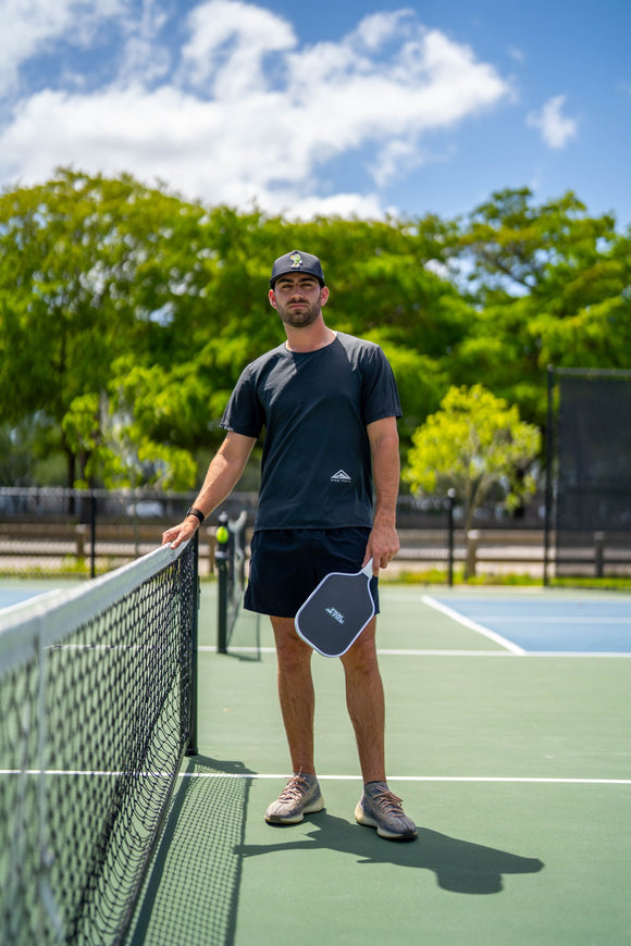 Player wearing a TMP cap and athletic gear standing next to a pickleball net, holding the TMP Stealth Pickleball Paddle by his side, trees and sky behind