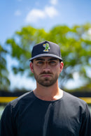 Close-up portrait of a player wearing a TMP cap featuring the pickle mascot character, with blurred trees and sky in the background