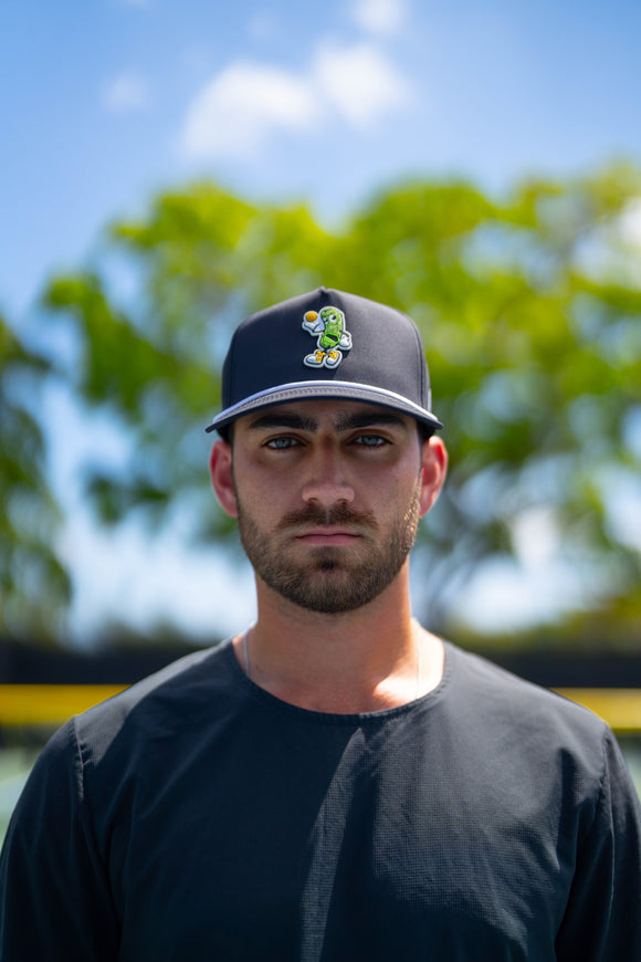 Close-up portrait of a player wearing a TMP cap featuring the pickle mascot character, with blurred trees and sky in the background