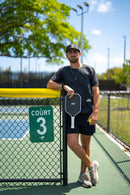 Player in a TMP cap and athletic gear leaning against a Court 3 chain-link fence, holding the TMP Stealth Pickleball Paddle and a yellow pickleball