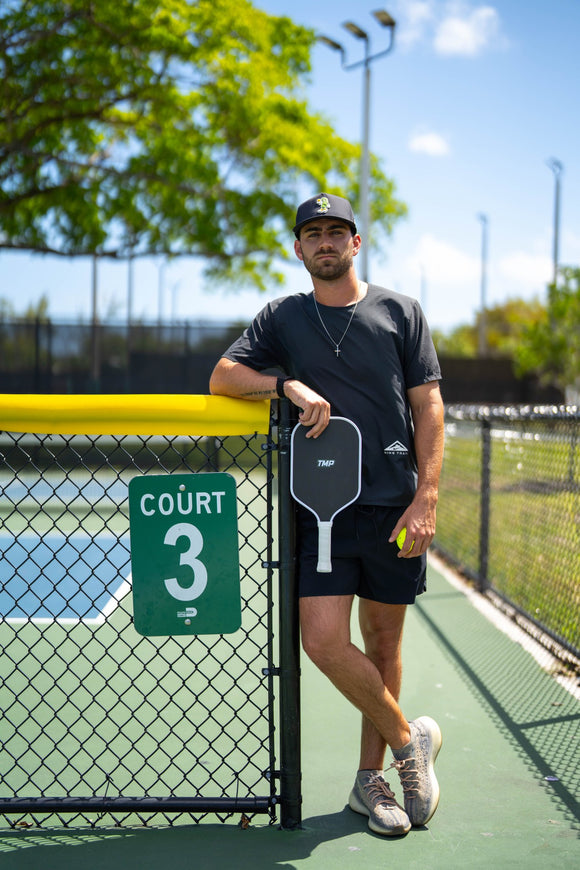 Player in a TMP cap and athletic gear leaning against a Court 3 chain-link fence, holding the TMP Stealth Pickleball Paddle and a yellow pickleball